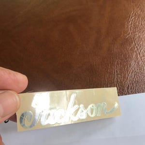 Puede incluir: Una pegatina met&aacute;lica plateada con la palabra "Jackson" escrita en cursiva sobre un fondo beige.