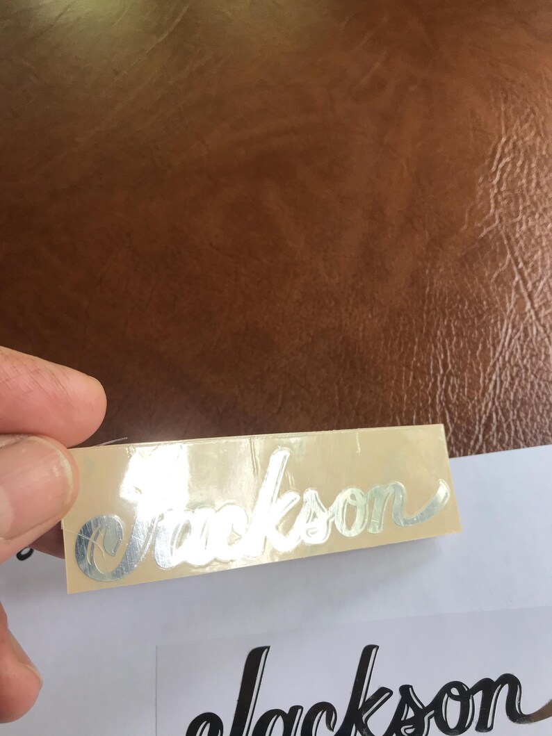 Puede incluir: Una pegatina met&aacute;lica plateada con la palabra "Jackson" escrita en cursiva sobre un fondo beige.