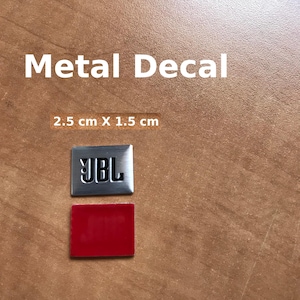 May include: A silver metal decal with the text "JBL" in black lettering. The decal is 2.5 cm by 1.5 cm and is on a brown wooden surface. The decal is accompanied by a red adhesive backing.