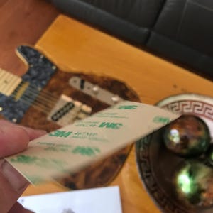 May include: A close-up of a brown and tan guitar with a 3M adhesive tape on top of it. The guitar has a black pickguard and a gold-colored bridge.