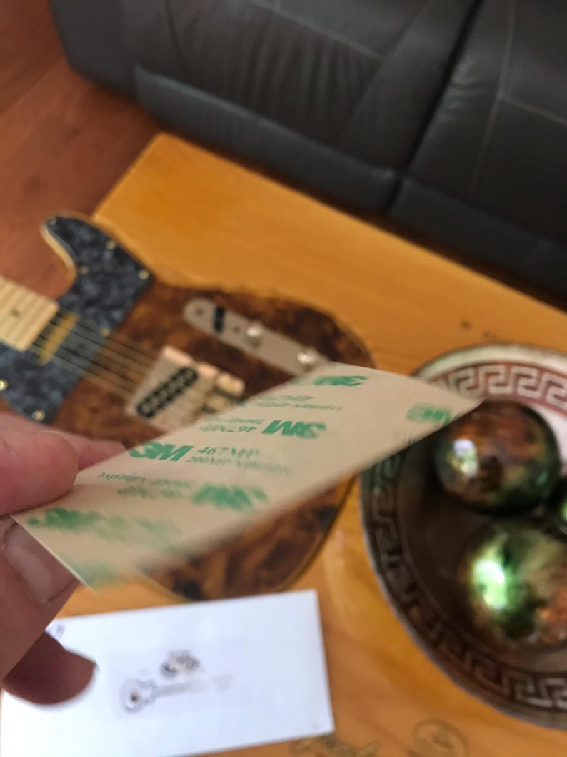May include: A close-up of a brown and tan guitar with a 3M adhesive tape on top of it. The guitar has a black pickguard and a gold-colored bridge.