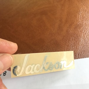 Puede incluir: Una pegatina plateada con la palabra "Jackson" escrita en cursiva sobre un fondo beige.