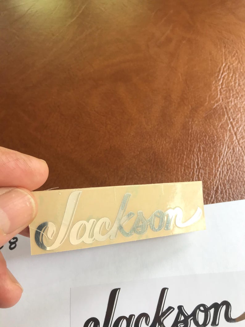 Puede incluir: Una pegatina plateada con la palabra "Jackson" escrita en cursiva sobre un fondo beige.