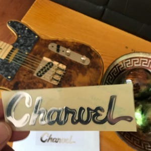 May include: A silver Charvel guitar logo sticker on a beige background. The sticker is slightly curved and has a glossy finish.