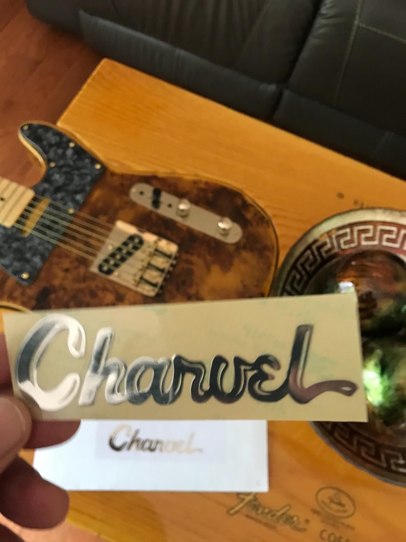 May include: A silver Charvel guitar logo sticker on a beige background. The sticker is slightly curved and has a glossy finish.