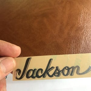 Puede incluir: Primer plano de una pegatina negra con el logotipo "Jackson" sobre un fondo beige.