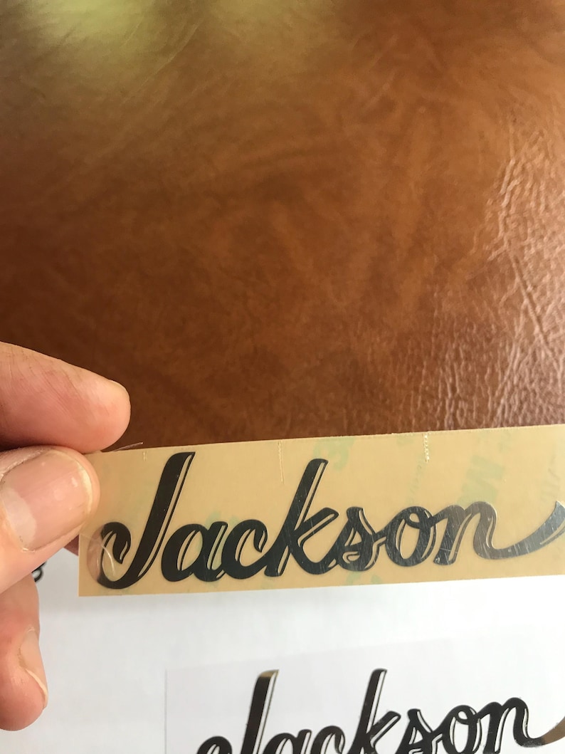 Puede incluir: Primer plano de una pegatina negra con el logotipo "Jackson" sobre un fondo beige.