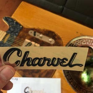 May include: A black vinyl sticker with the word "Charvel" in a stylized font. The sticker is on a clear background and is being held by a hand.