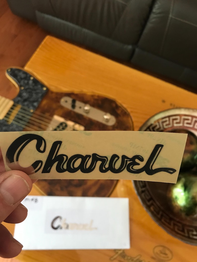 May include: A black vinyl sticker with the word "Charvel" in a stylized font. The sticker is on a clear background and is being held by a hand.