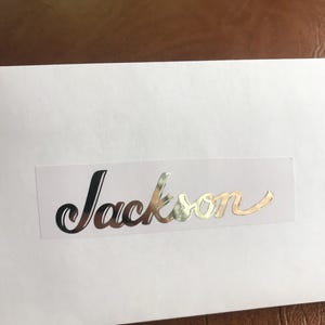 Puede incluir: Una tarjeta blanca con el nombre "Jackson" escrito en letras cursivas plateadas.
