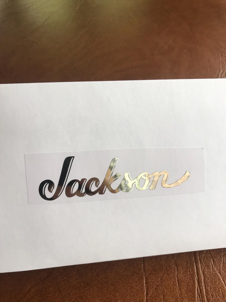 Puede incluir: Una tarjeta blanca con el nombre "Jackson" escrito en letras cursivas plateadas.
