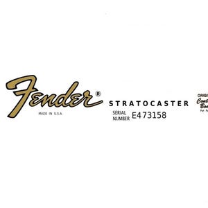 Puede incluir: Logotipo dorado de Fender, con la palabra "Stratocaster" y el número de serie E473158. El texto "Made in U.S.A." está debajo del logotipo. También son visibles las palabras "Original Contour Body".