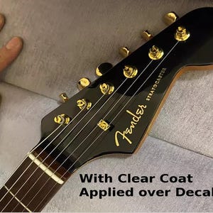 Puede incluir: Una guitarra el&eacute;ctrica Fender Stratocaster negra con clavijas doradas y una capa transparente aplicada sobre la calcoman&iacute;a. La guitarra tiene un diapas&oacute;n de palisandro y una golpeadora blanca.
