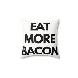 May include: A square throw pillow with a white fabric cover featuring the text EAT MORE BACON printed in bold black capital letters across the center. The pillow has a standard square shape with rounded corners and a smooth, clean surface finish.