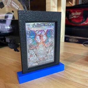 May include: A black card display stand with a blue base, showcasing a Pokemon trading card featuring Charizard EX with the text "Charizard EX 3300".