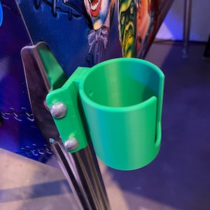 May include: A green 3D printed cup holder attached to a silver metal pole. The cup holder is designed to hold a beverage can or bottle.