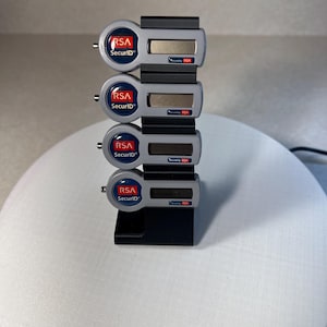 May include: Four gray RSA SecurID tokens are stacked on a black stand. Each token has a red RSA logo and a small screen.
