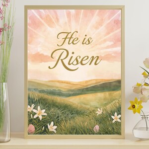 Könnte beinhalten: Gerahmter Aquarelldruck mit dem Text "He is Risen" in goldener Schrift. Das Kunstwerk zeigt einen Sonnenaufgang über sanften grünen Hügeln, weißen Lilien und Ostereiern. Der Rahmen ist hellgoldfarben.