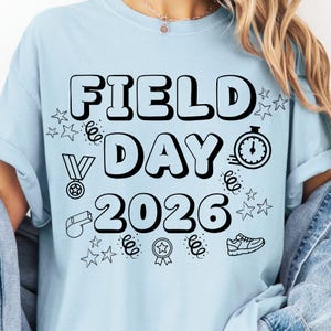 Può includere: T-shirt azzurra con la scritta "FIELD DAY 2026" in grandi lettere nere. La maglietta presenta varie illustrazioni nere di stelle, un cronometro, una medaglia, un fischietto, un nastro e una scarpa da corsa.