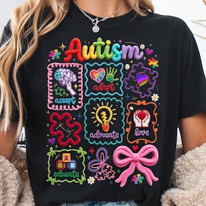 May include: Black t-shirt with colorful embroidered designs related to autism awareness. The word "Autism" is in rainbow colors. Other designs include a brain, heart, puzzle pieces, and the words "accept", "adapt", "advocate", "love", and "educate".