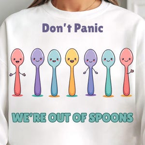 Könnte beinhalten: Weißes Sweatshirt mit dem Text "Don't Panic" über einer Reihe von sieben bunten Cartoon-Löffeln mit lächelnden Gesichtern. Unter den Löffeln steht der Text "WE'RE OUT OF SPOONS".