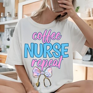 Könnte beinhalten: Ein weißes T-Shirt mit kurzen Ärmeln und dem Text "coffee NURSE repeat" in rosa und blauer Schrift. Unter dem Text befindet sich eine Grafik eines Stethoskops mit einer großen, schillernden, mehrfarbigen Schleife am Bruststück.