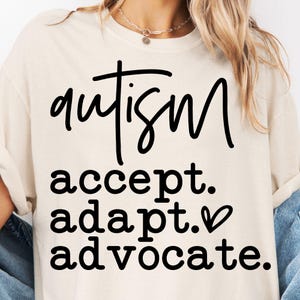 Könnte beinhalten: Cremefarbenes T-Shirt mit dem Wort "Autismus" in schwarzer, handschriftlicher Schrift. Darunter stehen die Worte "accept. adapt. advocate." in ähnlicher schwarzer Schrift, mit einem Herzsymbol neben "adapt."