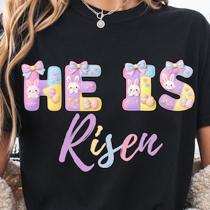 Könnte beinhalten: Schwarzes T-Shirt mit dem Schriftzug "HE IS Risen" in bunten Pastellfarben. Die Buchstaben "HE IS" sind mit Hasen- und Osterei-Designs verziert. Das Wort "Risen" ist in einer Schreibschrift gehalten.