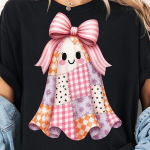 Könnte beinhalten: Ein schwarzes T-Shirt mit einer zentralen Grafik eines Geistes, der aus verschiedenen Patchwork-Stoffquadraten in orangefarbenen, rosa und violetten Mustern besteht, darunter Vichy-Karo, Sterne und Blumen, gekrönt von einer großen rosa-weißen Schleife.