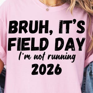 Può includere: T-shirt rosa chiaro con la scritta nera "BRUH, IT'S FIELD DAY I'm not running 2026" in un carattere invecchiato. La maglietta è in stile casual, adatta all'uso quotidiano.