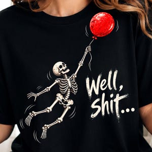 Könnte beinhalten: Ein schwarzes T-Shirt mit einer Grafik eines weißen Skeletts, das nach oben schwebt und einen einzelnen roten Luftballon hält. Rechts neben dem Skelett steht der Text "Well, shit..." in einer abgenutzten weißen Schrift mit Schatten.