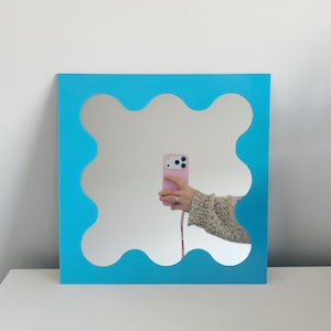 May include: Square mirror with a wavy, light blue frame. The mirror reflects a person holding a pink phone. The frame has a unique, decorative design, adding a modern touch to the wall decor.