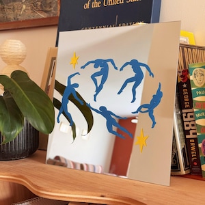 May include: A square mirror featuring five blue human-shaped silhouettes in various dancing poses and two yellow four-pointed stars. The mirror reflects the surrounding room, including a green plant and several books with visible text on their spines.