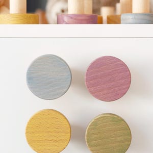 May include: Four round wooden cabinet knobs in pastel colors: light blue, pink, yellow, and green. The knobs are mounted on a white surface. The knobs are part of a set of wooden toys.