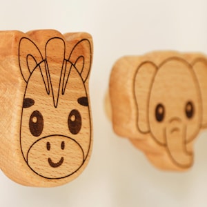 Könnte beinhalten: Holzschubladengriffe mit Tiergesichtern. Die Zebra- und Elefantenmotive sind in das Holz geschnitzt und haben schwarze Umrisse. Die Griffe sind hellbraun mit glatter Oberfläche, ideal für ein Kinderzimmer.