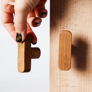 Può includere: Due pomelli per mobili in legno marrone chiaro. Uno a forma di T è tenuto in mano, l'altro è attaccato a una superficie in legno. I pomelli hanno una finitura liscia e naturale.