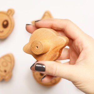Puede incluir: Juguetes de madera con tem&aacute;tica espacial, que incluyen un extraterrestre, un cohete y un OVNI. El OVNI se sostiene en una mano. Los juguetes son de color marr&oacute;n claro y tienen detalles grabados. El fondo es blanco.