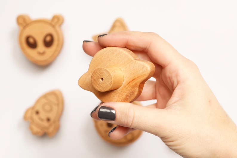 Puede incluir: Juguetes de madera con tem&aacute;tica espacial, que incluyen un extraterrestre, un cohete y un OVNI. El OVNI se sostiene en una mano. Los juguetes son de color marr&oacute;n claro y tienen detalles grabados. El fondo es blanco.