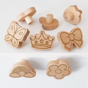 Könnte beinhalten: Eine Sammlung von Holzschrankknöpfen in verschiedenen Formen, darunter ein Schmetterling, eine Krone, eine Schleife, ein Pilz und eine Blume. Jeder Knopf ist aus hellem Holz mit geätzten Details gefertigt, ideal zum Dekorieren von Möbeln.
