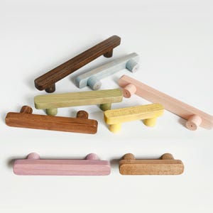 Op de afbeelding: Een verzameling van zeven houten ladehandgrepen in diverse kleuren en afwerkingen, waaronder donkerbruin, lichtbruin, saliegroen, lichtgeel, zachtroze en lichtblauw. Elke handgreep heeft een rechthoekig ontwerp met twee cilindrische bevestigingen.