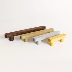 Peut inclure: Quatre poignées de placard en forme de T disposées en rangée, diminuant en longueur de gauche à droite. Les poignées présentent différentes finitions : bois brun foncé, bois beige clair, gris clair mat et bois peint en jaune vif.