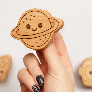 Puede incluir: Juguete de madera con forma de planeta con una cara sonriente y un anillo alrededor. El juguete est&aacute; sostenido por una mano, con un cohete y un juguete con forma de astronauta en el fondo. Los juguetes son de madera clara.