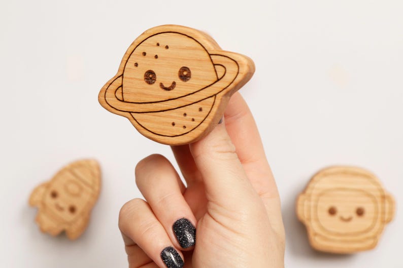 Puede incluir: Juguete de madera con forma de planeta con una cara sonriente y un anillo alrededor. El juguete est&aacute; sostenido por una mano, con un cohete y un juguete con forma de astronauta en el fondo. Los juguetes son de madera clara.
