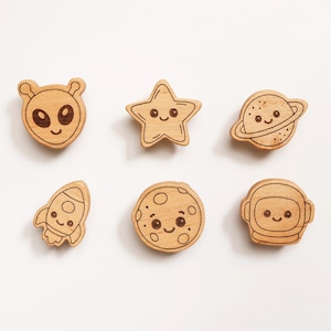 May include: Wooden space-themed embellishments featuring an alien, star, planet, rocket, moon, and astronaut. Each piece has a smiling face and is made of light-colored wood. Great for crafting and DIY projects.