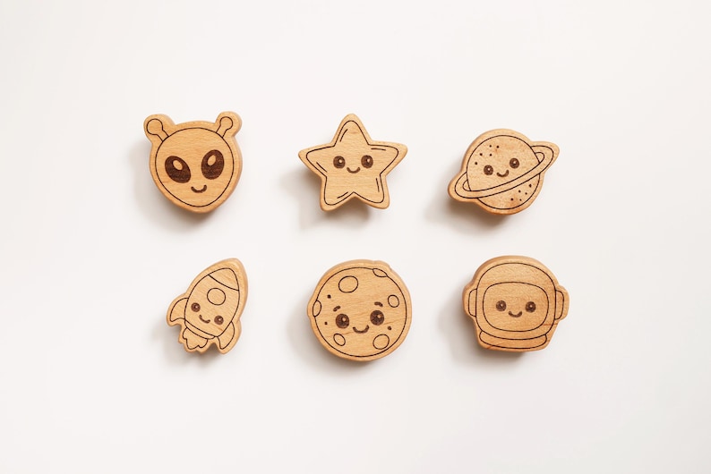 Puede incluir: Adornos de madera con tem&aacute;tica espacial, que representan un extraterrestre, una estrella, un planeta, un cohete, una luna y un astronauta. Cada pieza tiene una cara sonriente y es de madera clara. Para manualidades.