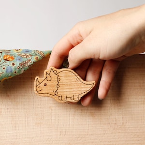 Könnte beinhalten: Ein Holzspielzeug in Form eines Triceratops mit geätzten Details. Der Dinosaurier ist hellbraun mit schwarzer Umrandung. Im Hintergrund ist ein Stoff mit floralem Muster zu sehen.