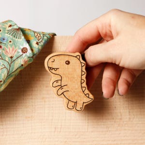 Puede incluir: Un juguete de madera con forma de dinosaurio con detalles de contorno negro. El juguete es de color marrón claro y está sostenido por una mano. En el fondo se ve una tela con estampado floral. El dinosaurio tiene una expresión amigable.