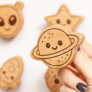 Puede incluir: Formas de madera con tem&aacute;tica espacial, incluyendo un alien&iacute;gena, una estrella, un cohete y un planeta con anillos. El planeta es sostenido por una mano con una u&ntilde;a de purpurina negra. Las formas tienen detalles grabados y caras sonrientes.