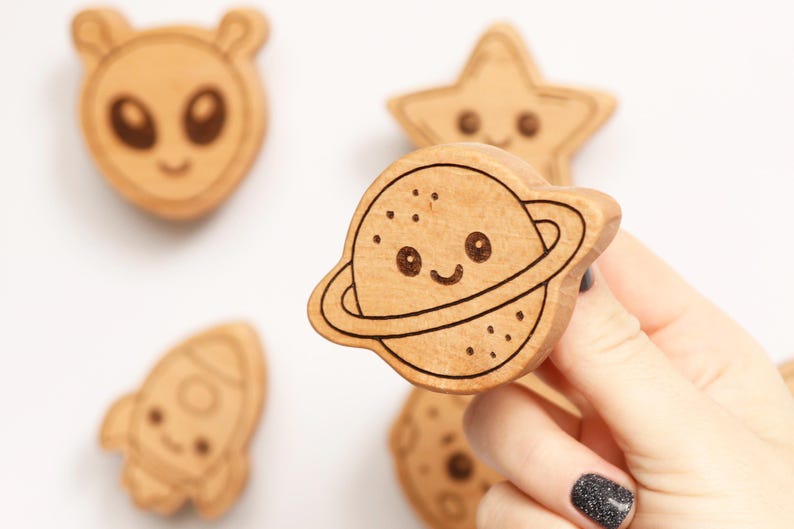 Puede incluir: Formas de madera con tem&aacute;tica espacial, incluyendo un alien&iacute;gena, una estrella, un cohete y un planeta con anillos. El planeta es sostenido por una mano con una u&ntilde;a de purpurina negra. Las formas tienen detalles grabados y caras sonrientes.