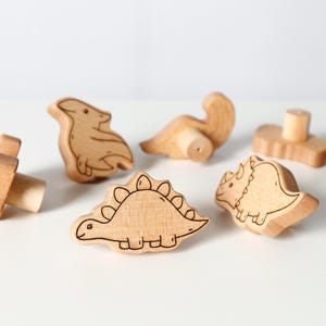 May include: A collection of wooden dinosaur toys, including a Stegosaurus, Triceratops, and Pterodactyl, with black outline details. The toys are made of light-colored wood and are arranged on a white surface.